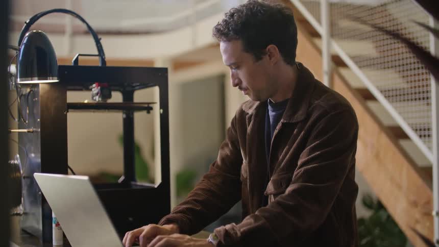 A man standing at a table in front of a laptop next to a manufacturing device as the man looks stressed