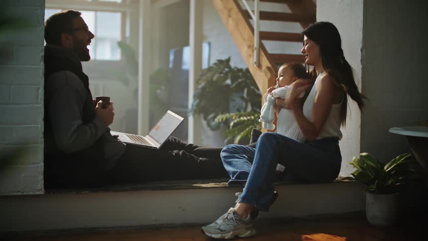 Mother with straight hair white shirt and jeans holds baby and father with beard glasses and hoodie uses laptop