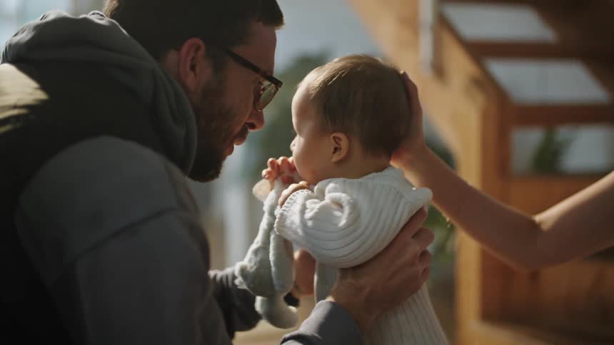 A man wearing glasses holding up a baby smiling and talking to him while a person puts their hand on the babies head