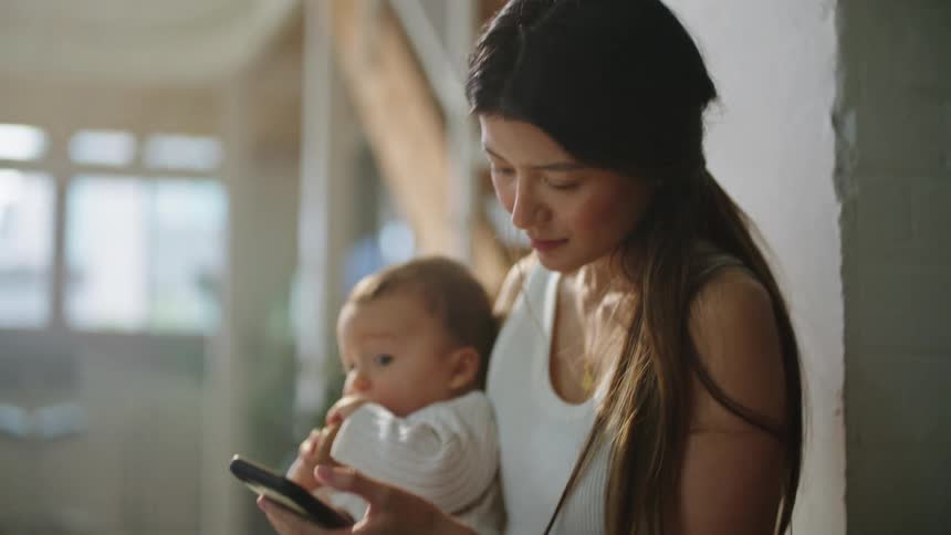 A woman uses a smartphone with one hand while holding a baby with her other arm
