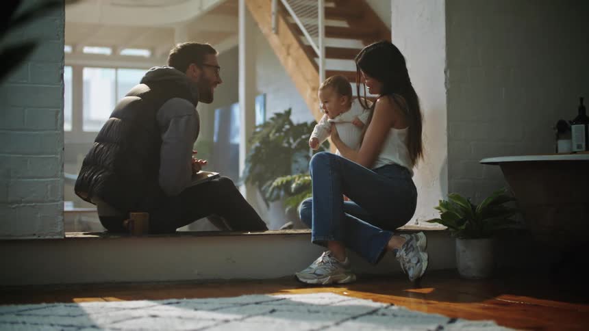 Mother with white shirt and father with glasses beard vest and hoodie play with baby at home