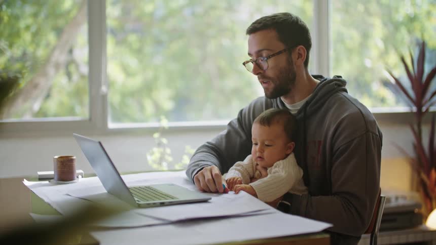 Baby sits on fathers lap and grabs papers and highlighter while dad participates in online work meeting through laptop