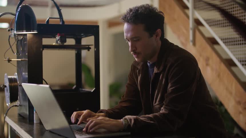 A man using a tool in an automatic robot like device before turning to type on his laptop