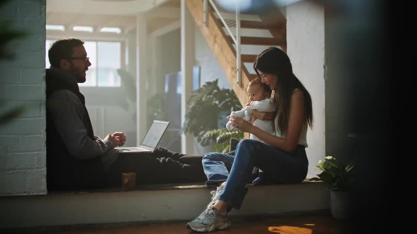A man works on his laptop while his wife holds their baby