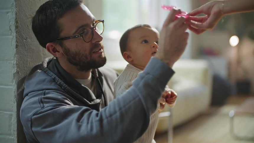 A man is holding his baby as his wife hands him a pacifier