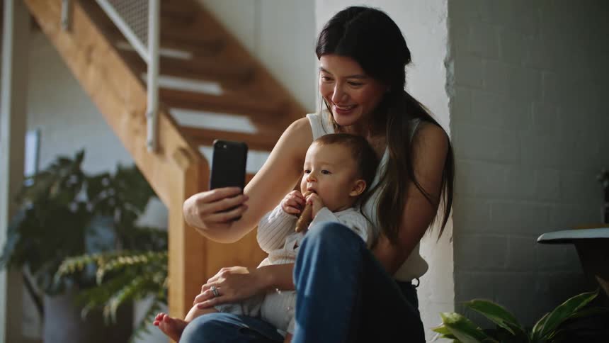 A woman takes a selfie with her baby while sitting on stairs at home