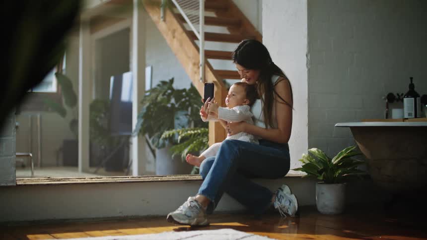 A mother holding her baby while they sit on a large bay window sill while the mother holds her cellphone in front of the baby