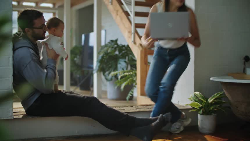 A man holds his baby while his wife walks away holding a laptop