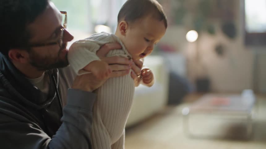 Father with beard glasses and gray hoodie sitting on floor holds baby at home