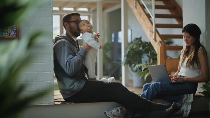 A woman is sitting with a computer in her lap as she types and talks to her husband as he holds their baby