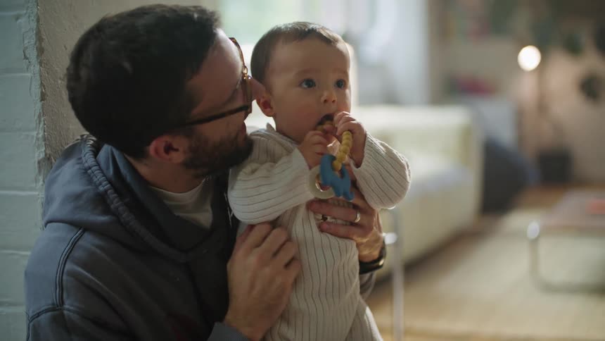 A father wearing glasses holding his baby son as another person hands him a toy for the child