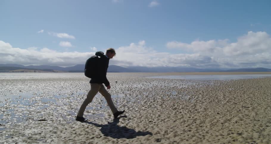 A man wearing a backpack walking across wet sand with a mountain landscape in the distance