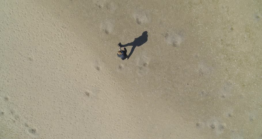 A man wearing a backpack and a jacket is walking across the desert as his shadow walks next to him
