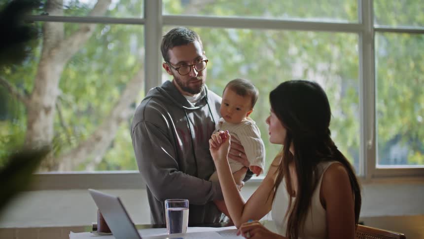 A father holds his baby son while he talks to his wife sitting at a table in front of a laptop and papers