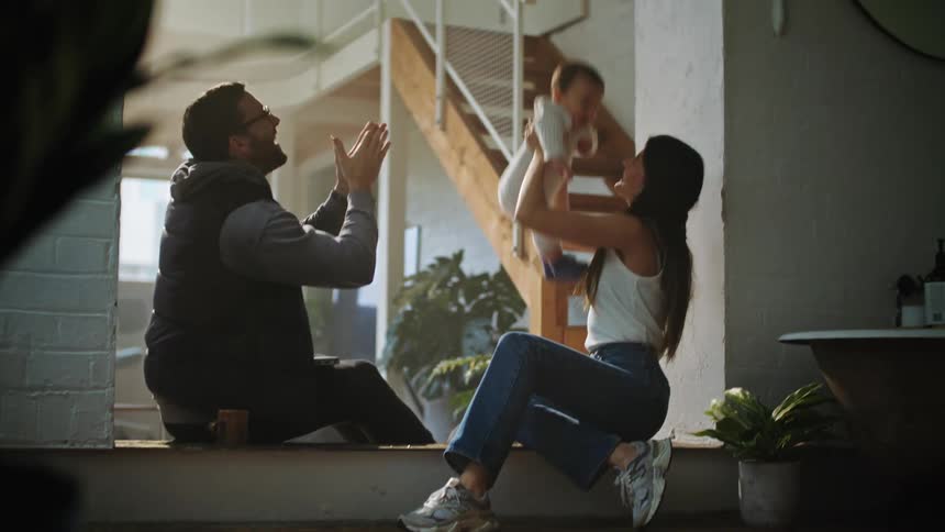 Father with beard and black jacket and pants sits on floor with mother wearing jeans and white shirt and toddler boy