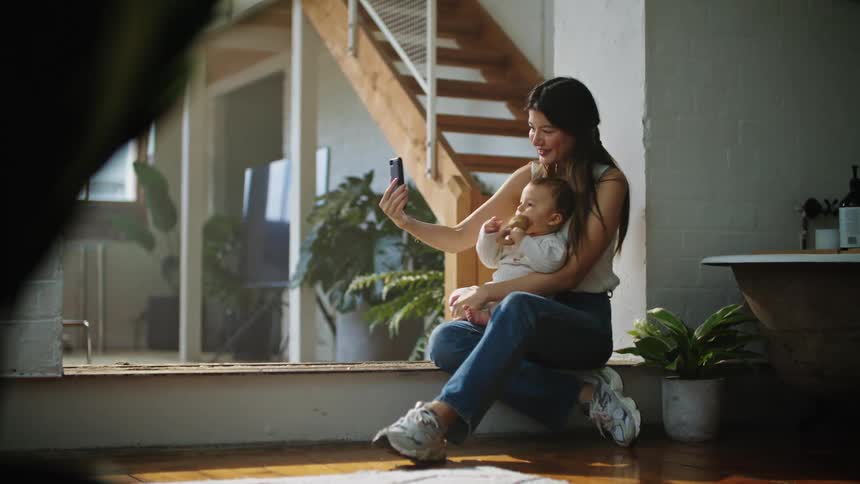 Mother with white shirt and jeans and shoes holds baby while taking selfie with smartphone