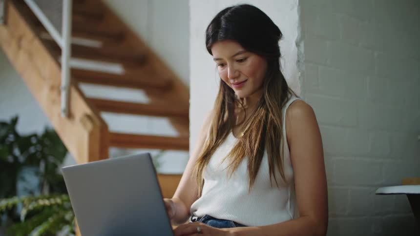 Woman leaning against brick wall and working on laptop looks up briefly with a smile