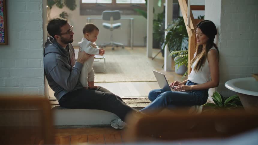Mother using laptop sits against wall across from father holding baby