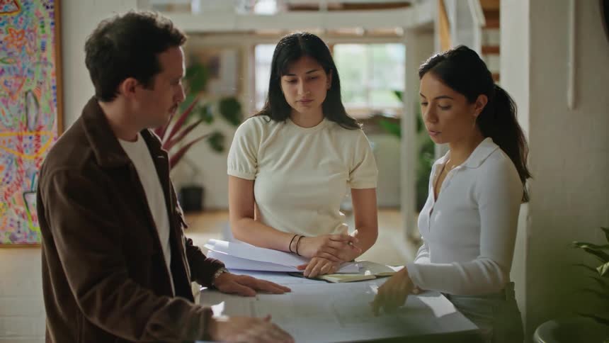 Two women and a man standing around a table looking at plans on a paper and conversing
