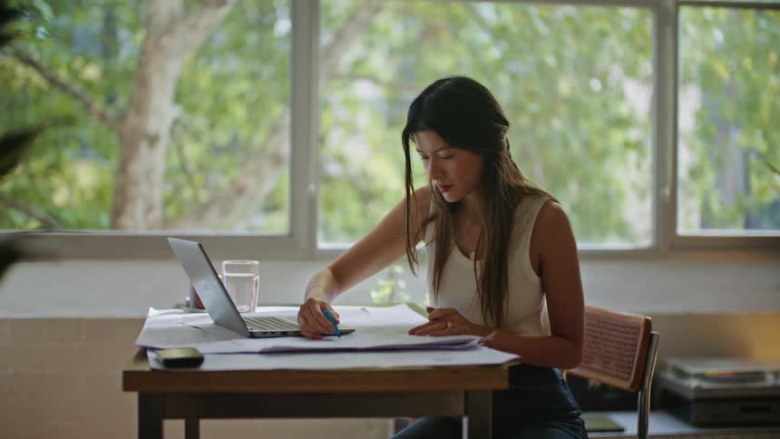 A woman sitting at a desk with papers on it a laptop in front of her and her cellphone on the corner