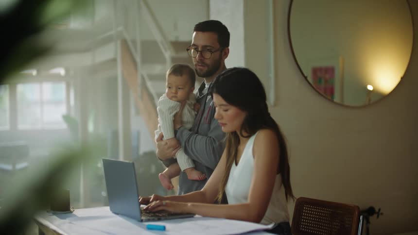 Father with glasses beard and gray hoodie holds baby while mom in white shirt working on laptop