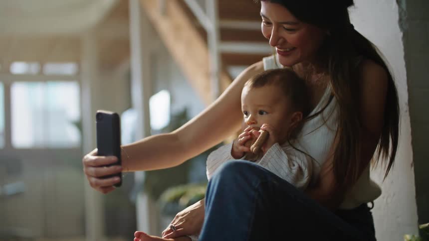 Mother uses smartphone to take video of baby she is holding while sitting near brick wall in home