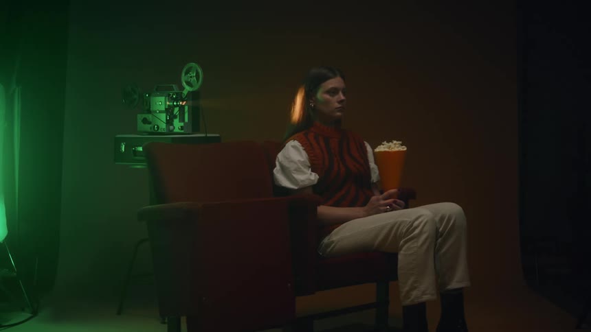 Woman with straight hair red sweater and beige pants watches movie holding popcorn while cinema camera running