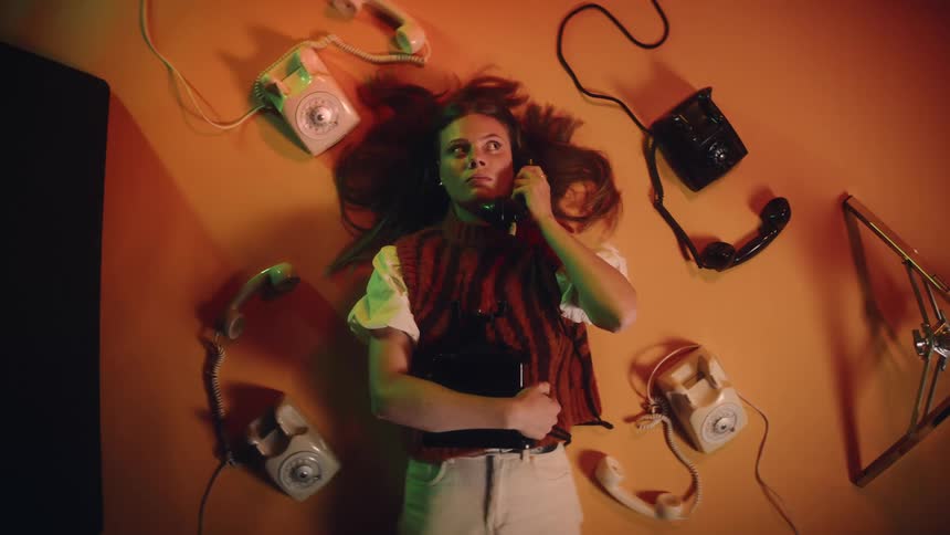 A woman lies near vintage telephones and puts a receiver to her ear to listen for something