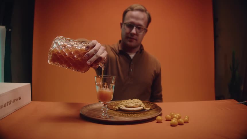 A man sitting at a table pouring a drink into his glass with food in front of him and an orange back drop