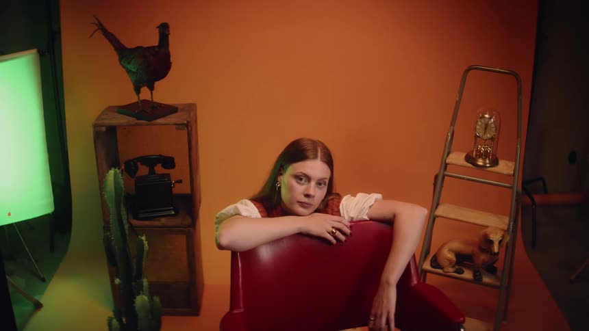 A woman crouched down behind a red chair on a photo set with an orange backdrop and decor on either side of her
