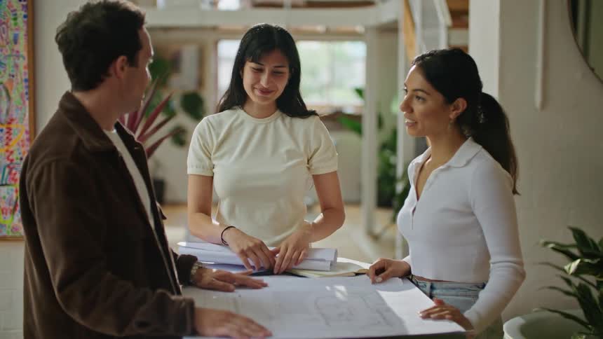 Man with mustache and brown jacket talks with women wearing white shirt working on plans at the office