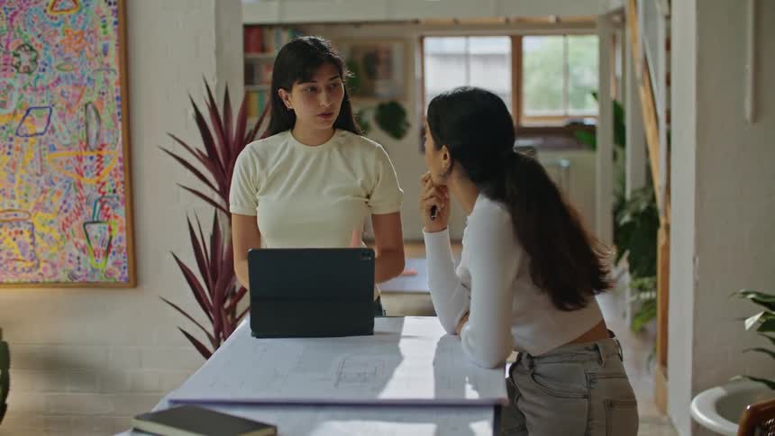 A woman standing in front of a laptop talking to another woman sitting at the same table while she looks at papers