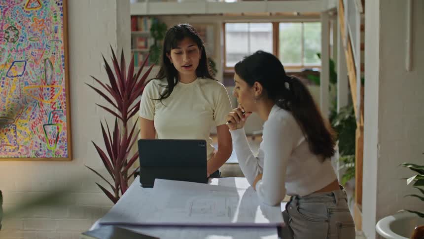 Two women one standing in front of a laptop while the other sits at the table looking over papers