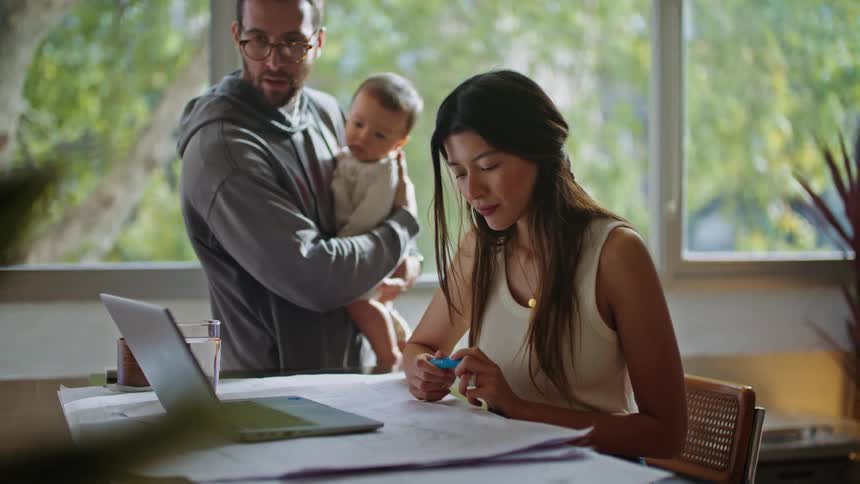 A man holding a baby standing next to his wife sitting at a desk while he points to what she is looking at and they converse about it