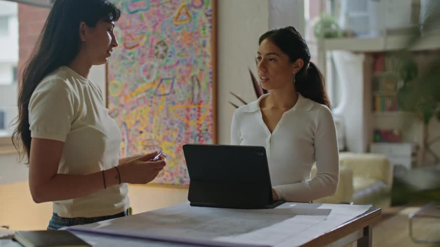 Two women discussing a design with laptop and blueprint on drafting table in office