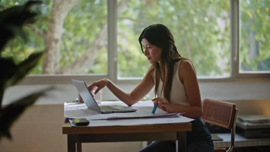 A woman sitting at her desk looking at and using a laptop while holding a blue pen