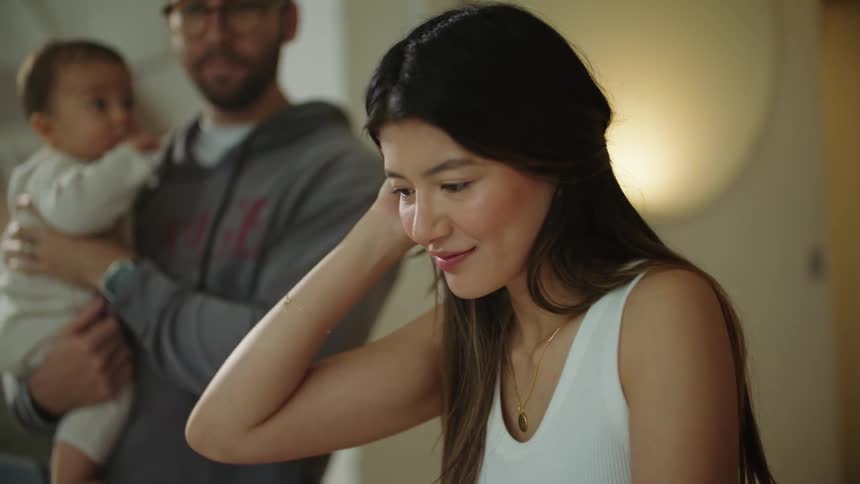 Woman with brown hair white shirt and necklace works on laptop while father rocking baby son in his arms