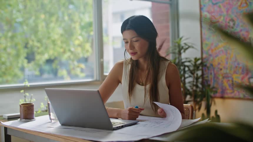 A woman sitting at her desk in front of a laptop looking and talking to it while looking at papers on her desk