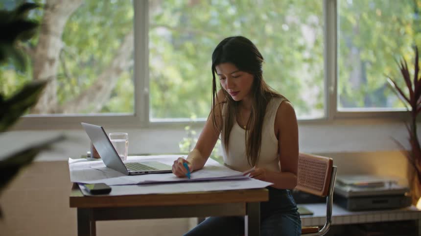 A woman is sitting at a desk working on a laptop computer and writing on plans