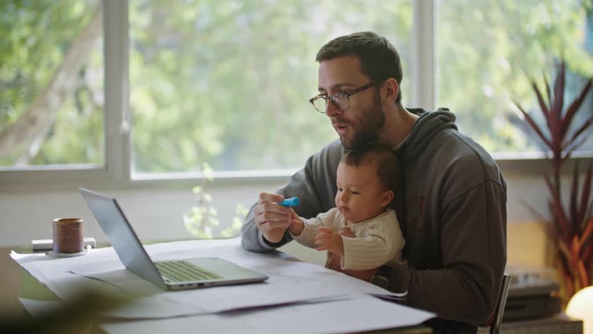 A man tries to work on his laptop with his baby in his lap