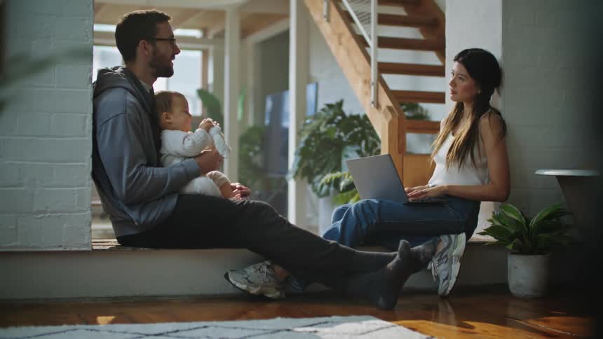 A woman is sitting with a computer in her lap as she types and talks to her husband as he holds their baby