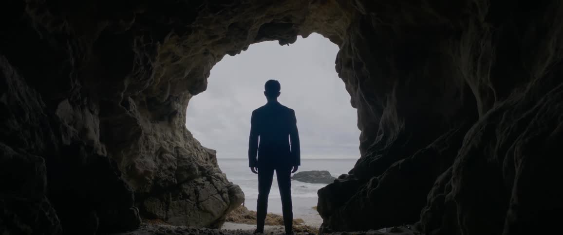A man stands near the opening of a cave by the beach