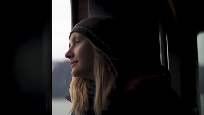 Blonde woman with gray wooly hat and black coat looks out the window smiling