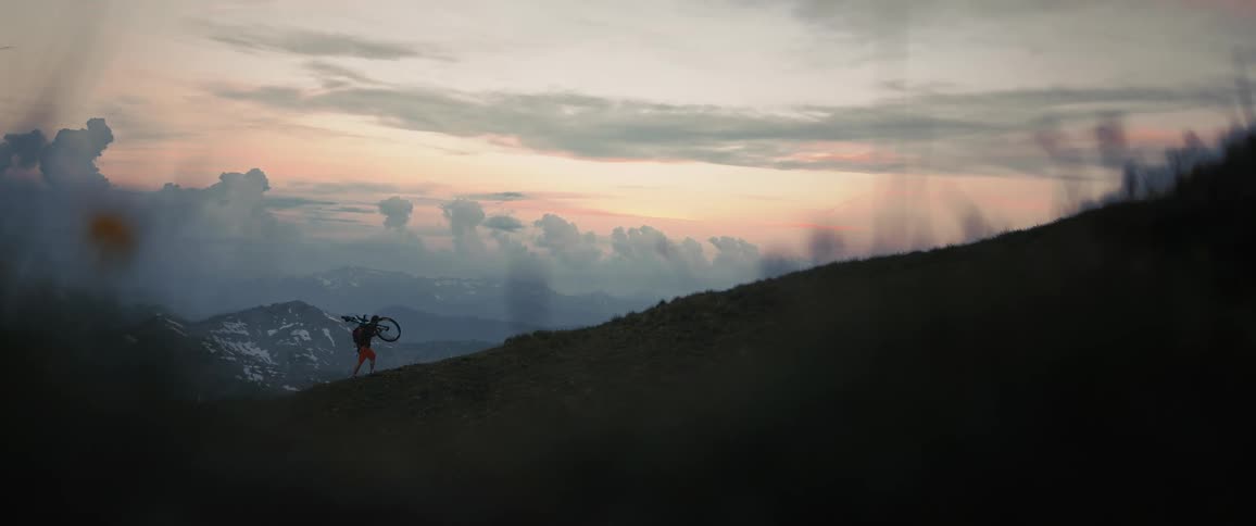 A person carries a bicycle on their shoulders while walking up a mountain slope in the evening