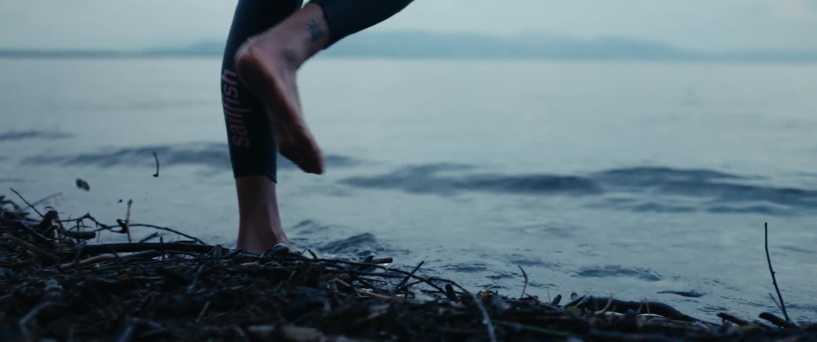 Person wearing wet suit walks barefoot into water as waves roll toward shore on foggy day