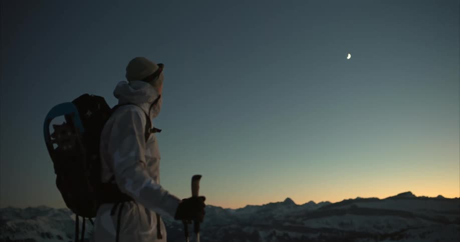 Man with hat coat backpack and trekking poles hikes in the snowy mountains at dusk