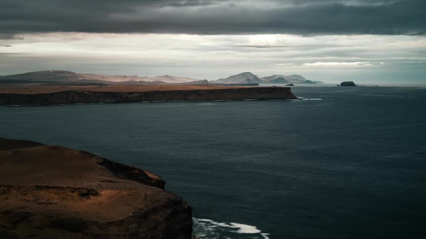 Sped up view of body of water moving along against rocky bank with mountains in the background and gray cloud cover