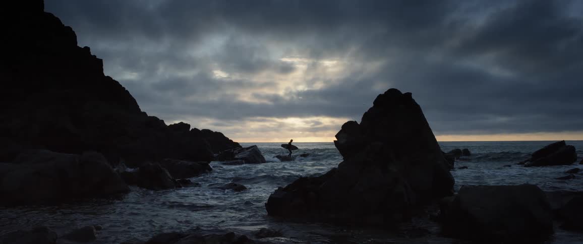 A surfer watches the waves while standing with a surfboard under their arm