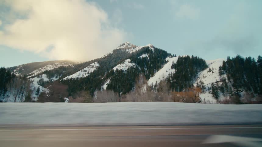 A view of a snowcovered mountain peak and green trees while moving down a nearby road