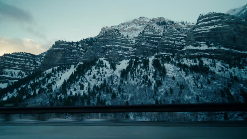 A shot from a nearby road of snowy mountain cliffs above a forest of pine trees in the day